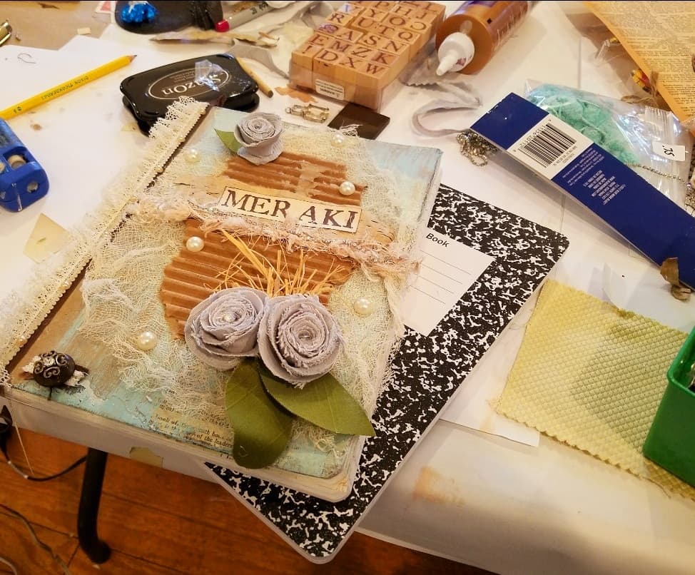 A decorated journal with floral embellishments and the word "MERAKI" on the cover, surrounded by crafting materials on a table.