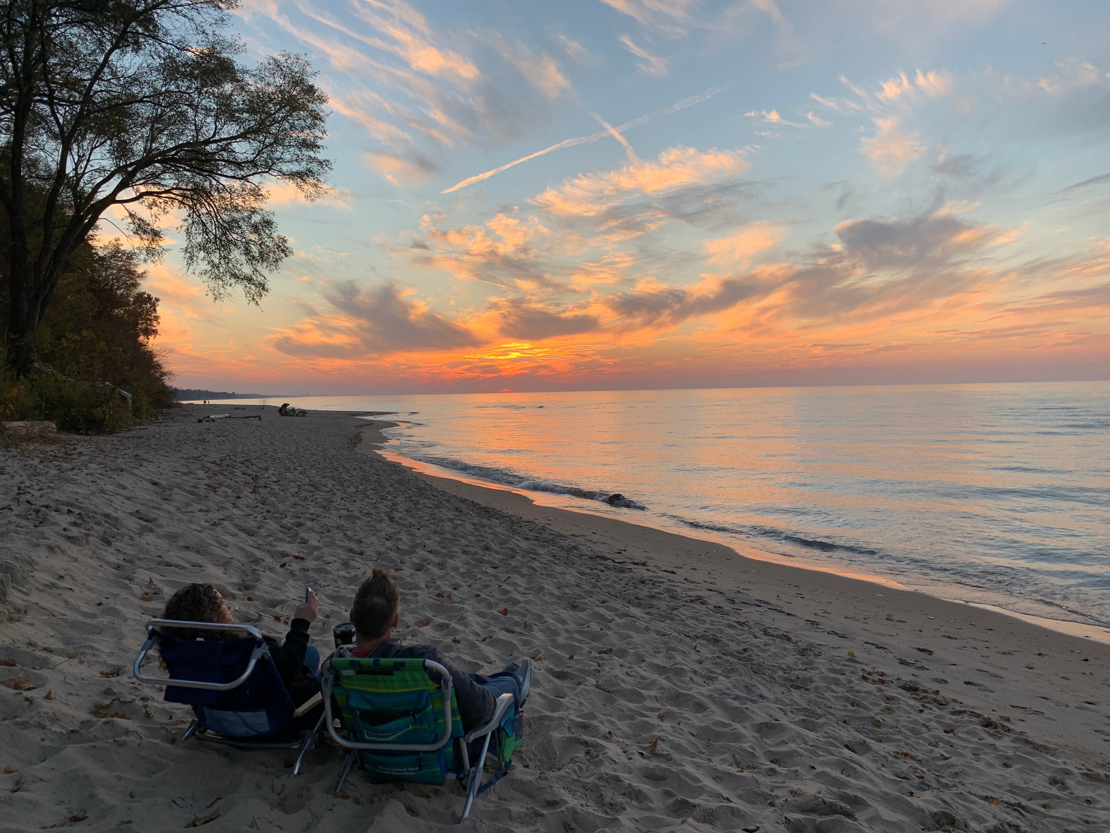 Two people sit on beach chairs, watching a colorful sunset over the calm water.