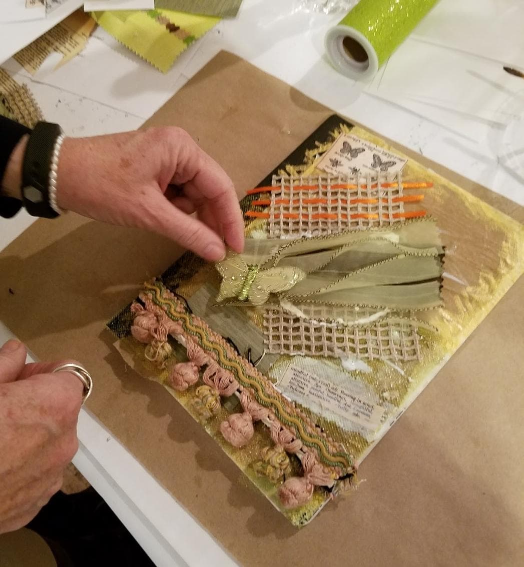A hand is placing a decorative butterfly onto a textured, mixed media art piece featuring ribbons, lace, and paper.
