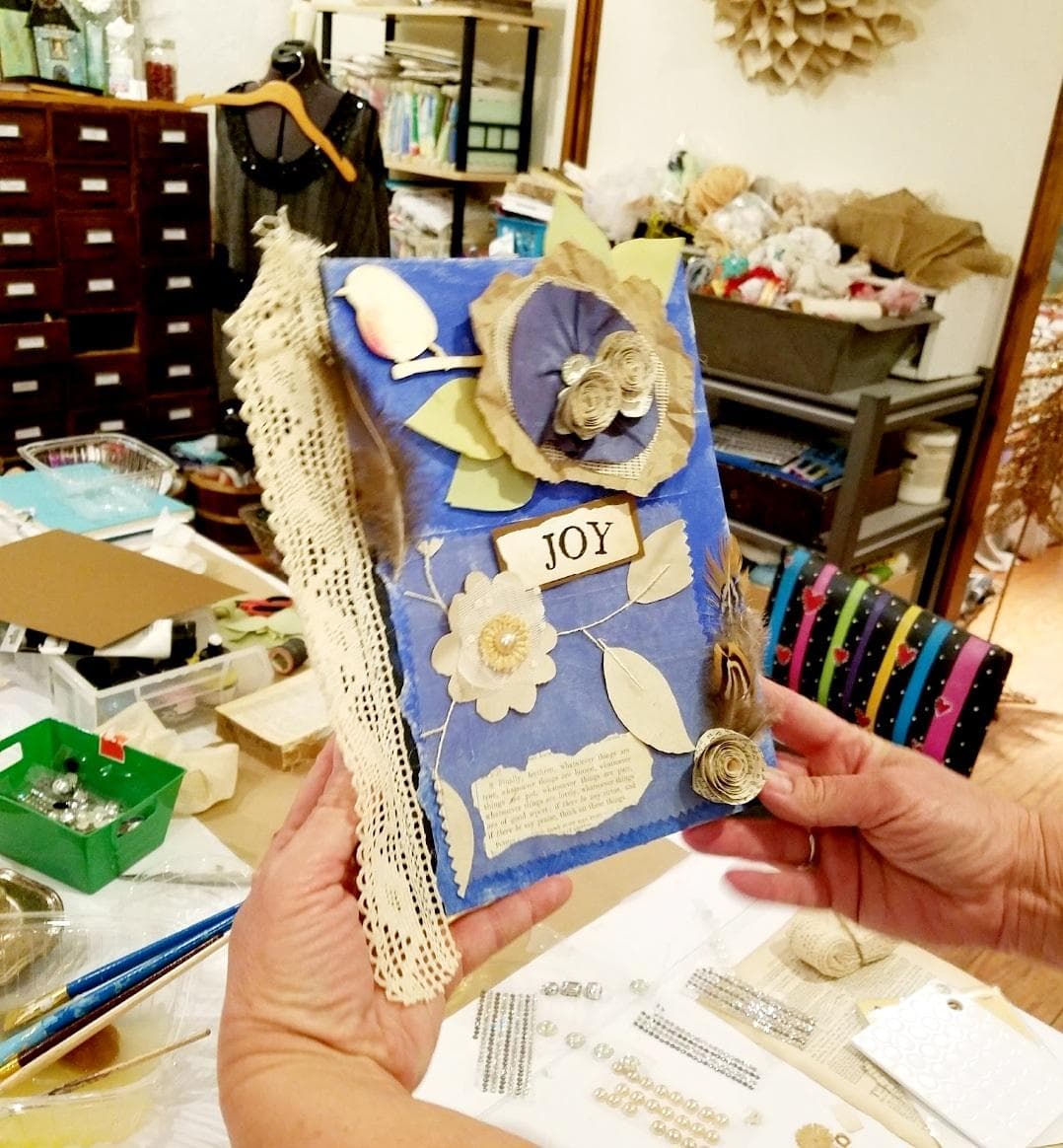 A person holds a decorated blue book featuring floral elements and the word "JOY" on its cover, surrounded by crafting materials.