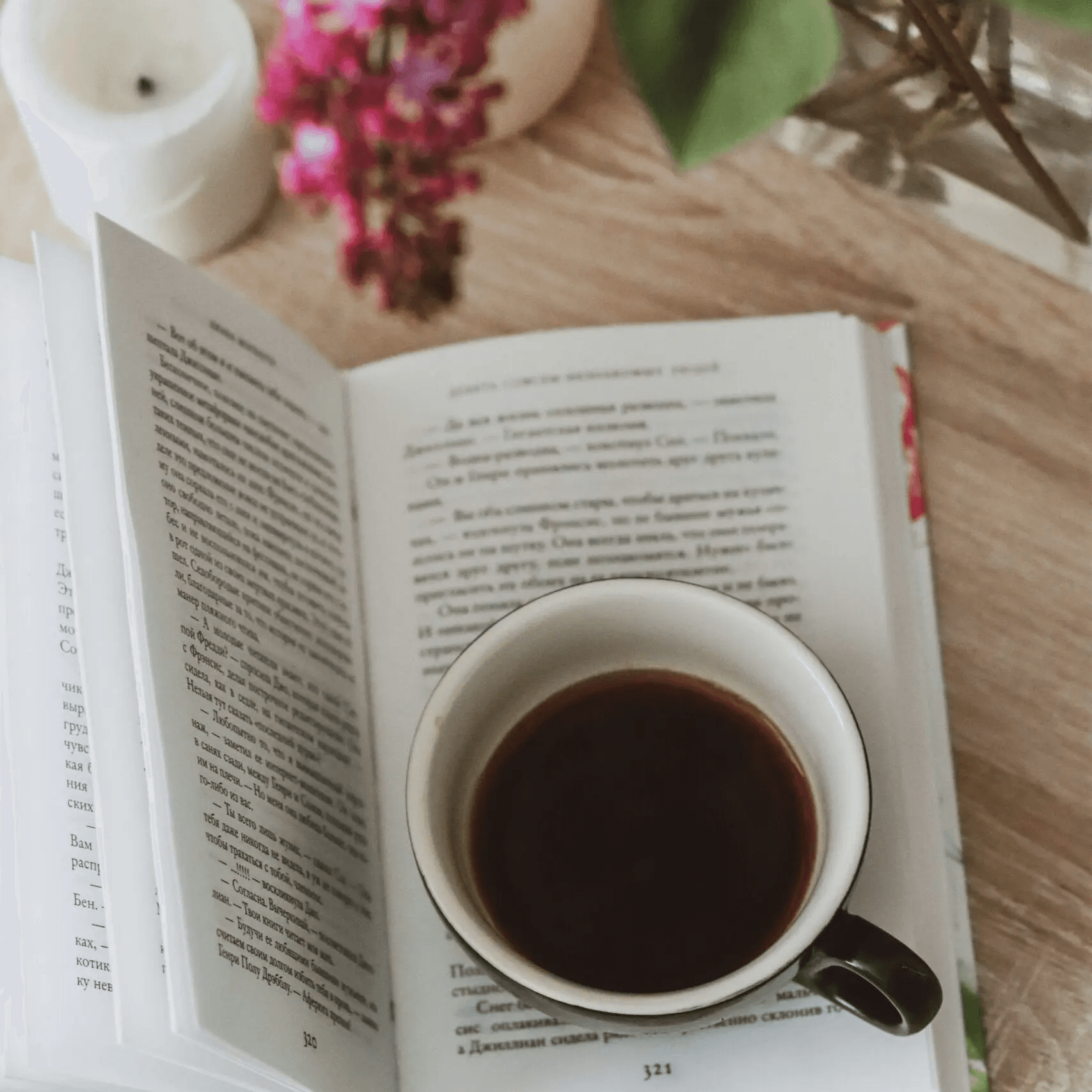 A coffee cup placed on an open book beside flowers and a candle.