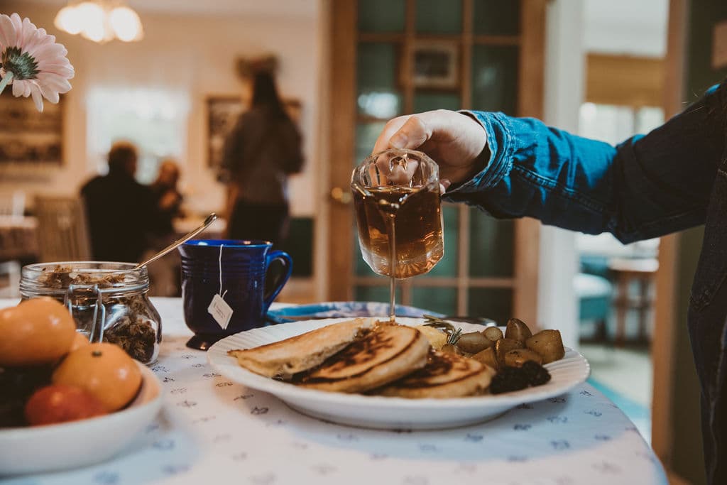 A hand pours tea over a plate of pancakes and fruit on a dining table.